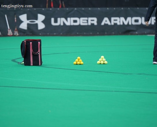 A sports field features a black and pink sports bag, two pyramids of yellow balls, and a portion of a person in dark athletic gear. The background displays a banner with the Under Armour logo.