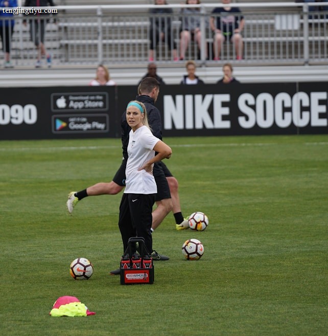 A person in sports attire stands on a soccer field, surrounded by several soccer balls and a carrier with bottles. The field is bordered by advertisements for Nike Soccer and app stores. Several people sit in bleachers in the background.