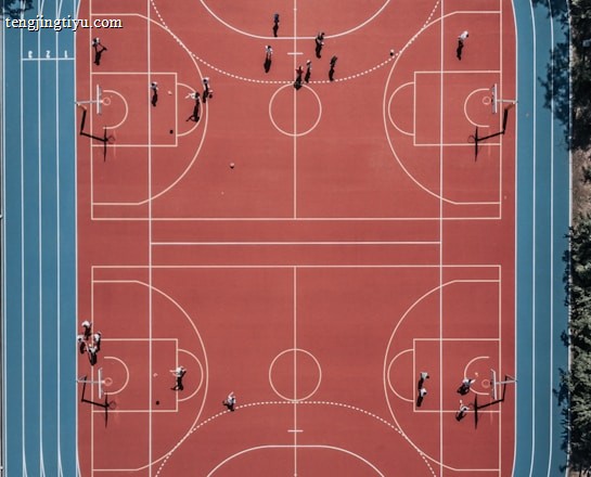 An aerial view of a large, vibrant sports complex featuring a red basketball court with multiple hoops and a surrounding blue running track. Several people are scattered across the area, engaged in various activities.