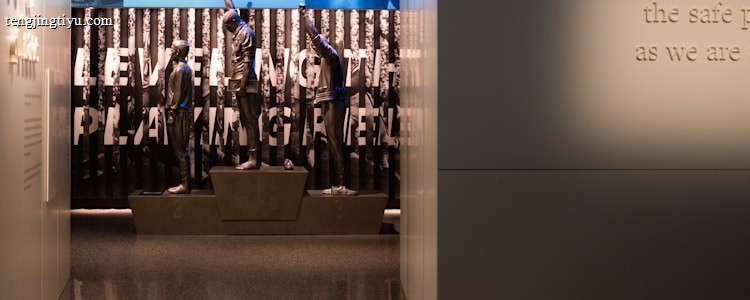 A dimly lit display features statues of athletes standing on a podium. They are positioned in front of a backdrop with the words 'LEVELING THE PLAYING FIELD' in bold letters. On the wall to the right, a softly lit quote is partially visible. The overall ambiance suggests a museum or exhibition setting.