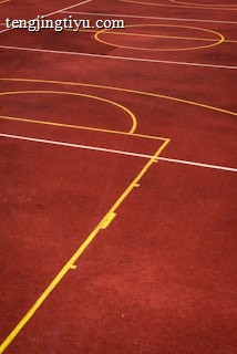 A sports court with a reddish-brown surface and bright yellow and white lines. The lines form geometric shapes, including rectangles and circles, suggesting markings for games like basketball or netball.