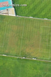 An aerial view of a sports complex featuring multiple green grass fields, including football pitches with marked goalposts. Adjacent to the fields is a blue-roofed building and a smaller area with a tennis or basketball court. Surrounding the complex, there's a road lined with trees and a dirt area with sparse vegetation.