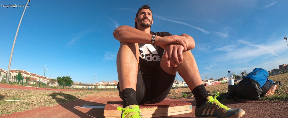 A person wearing a black athletic outfit is seated on a wooden platform in an outdoor sports field. The individual is relaxed, with arms resting on knees. The field is expansive with a clear blue sky and scattered clouds above. A brightly colored running shoe and athletic gear are visible, along with a water bottle and a blue backpack placed beside the person. Lampposts and residential buildings can be seen in the background.