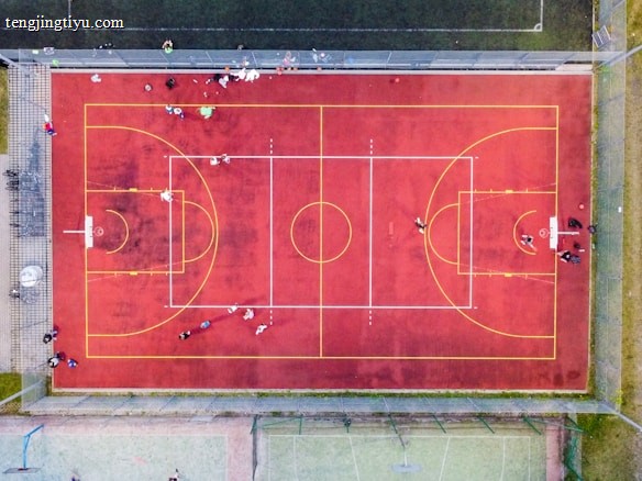 An aerial view of a sports court with a vibrant red surface and yellow lines marking different sections for basketball and soccer. Several people are present on the court, some engaged in activities, while others are standing along the perimeter. The court is enclosed by a metal fence and surrounded by grass on all sides.