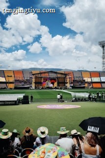 A sports stadium with a large stage set up on the field, hosting an outdoor event. Spectators wearing colorful hats sit in the foreground. The seating areas display vibrant colors, and a clear blue sky with scattered clouds is visible. Large screens display graphics related to the event.