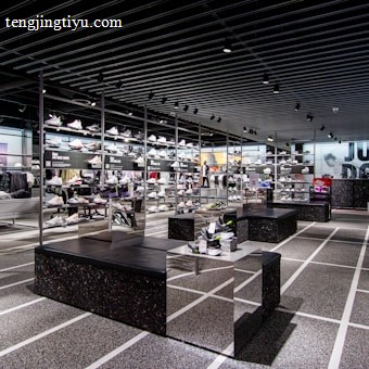 A modern sneaker store with sleek black shelving displaying various athletic shoes and sports apparel. Central seating areas and spotlights provide a contemporary retail ambiance. The signage prominently features a famous sportswear slogan in the background.