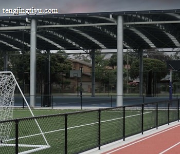 An outdoor sports facility featuring a running track and a soccer goal. The track is red, surrounded by a black metal fence, and the field is covered with artificial green turf. In the background, a basketball court is visible under a covered roof with metal supports. The sky is overcast, adding a moody atmosphere.