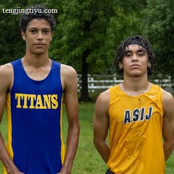Two young athletes wearing sleeveless sports tops stand on a grassy field. The athlete on the left is wearing a blue top with the word 'TITANS' while the one on the right wears a yellow top with 'ASIJ'. The background features green trees and a white fence, with a bench visible on the right.