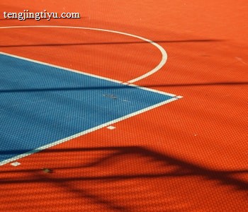 A vibrant sports court with sections of red and blue, featuring crisp white lines marking the boundaries. Shadows from a structure create geometric patterns across the surface, adding depth and interest to the scene.